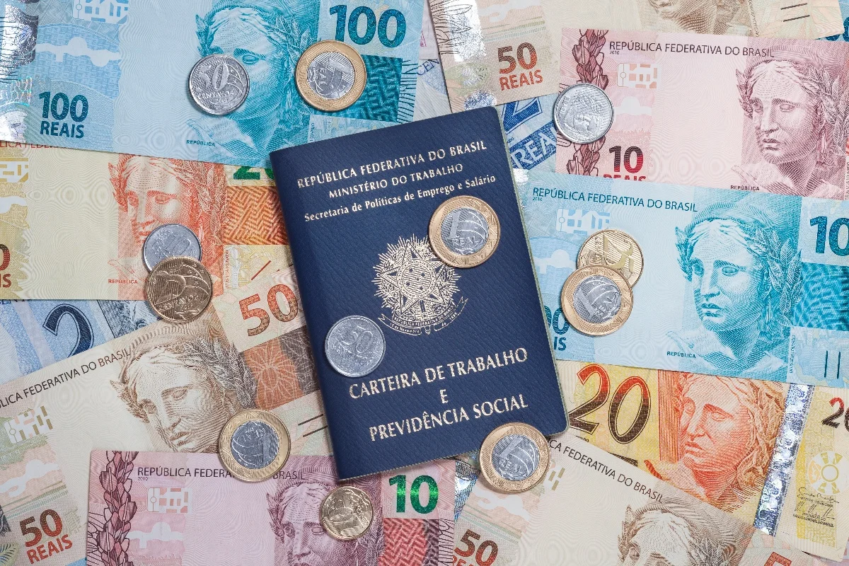 Foto colorida de notas de dinheiro Real e moedas, além de uma Carteira de Trabalho